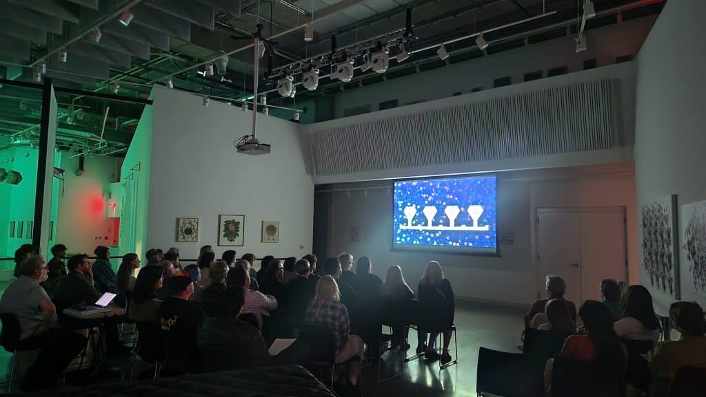 An arts gallery space is packed with people for a digital screening on a projection screen at the front of the room. The crowd all faces forward at the animated blue and white images on the screen. The far side of the galleries behind the attendees is mood-lit in a neon green.