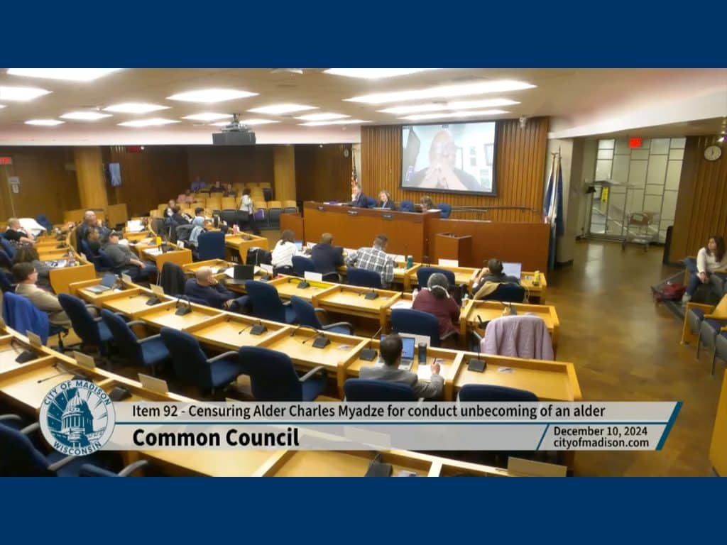 A screenshot from a video feed of a Madison Common Council meeting shows a large meeting chamber from the back, with various people sitting at desks arranged in a semicircle and facing a raised platform on which the Mayor and City Attorney sit. A chyron on the bottom reads "Item 92: Censuring Alder Charles Myadze for conduct unbecoming of an alder."