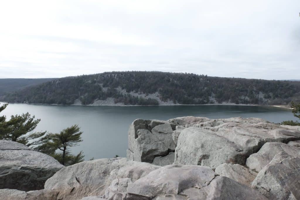A landscape photograph shows a lake with greenish water from atop a cliffside. A large cropping of trees appears on the opposite part of the lake. The sky is nearly a clear off-white color, and the general color palette of the image is muted on a late fall day.