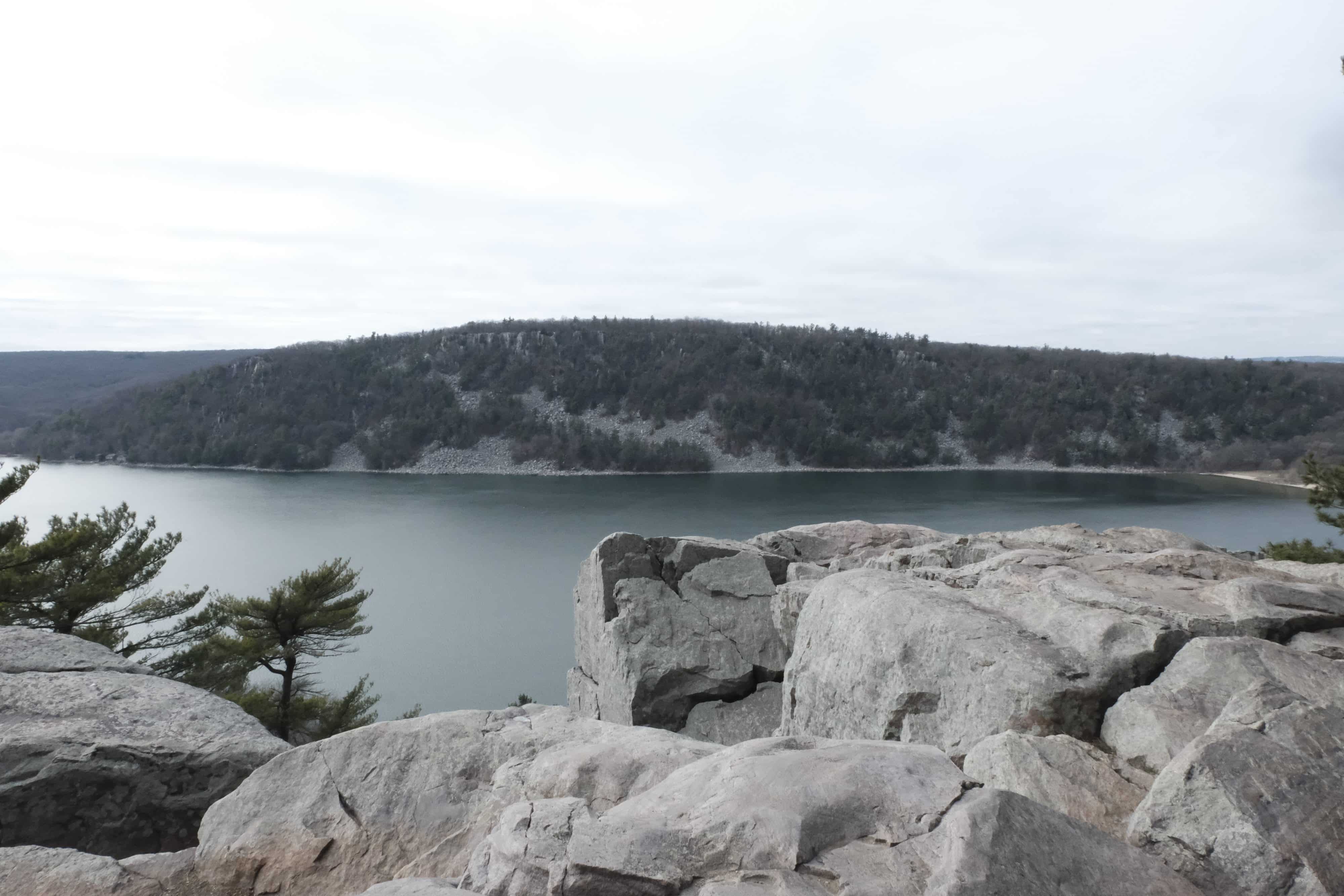 A landscape photograph shows a lake with greenish water from atop a cliffside. A large cropping of trees appears on the opposite part of the lake. The sky is nearly a clear off-white color, and the general color palette of the image is muted on a late fall day.