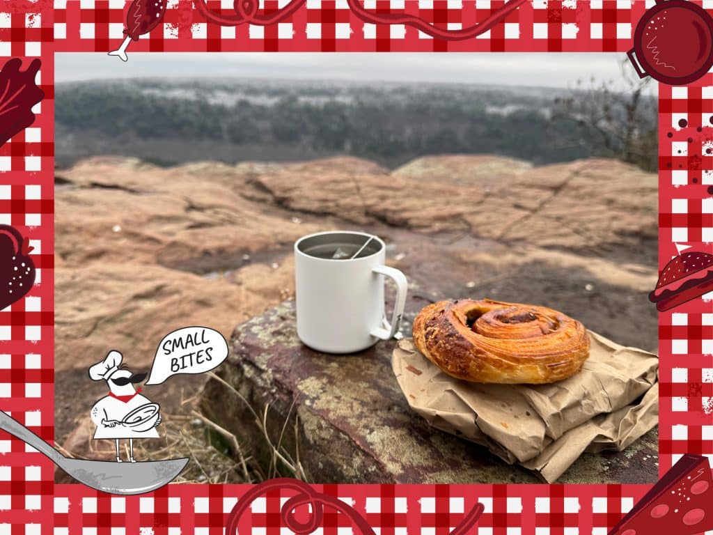 A winter photo atop a cliffside shows an expansive landscape of trees on the horizon. A flakey pastry and white mug with a hot beverage rest on a rock in the foreground. The photo is framed with an illustrated tablecloth with food items including pasta, cheese, a hamburger and more frame the photo. In the lower left corner, a small chef, with a comically large mustache, stands on a spoon and says, "Small Bites."