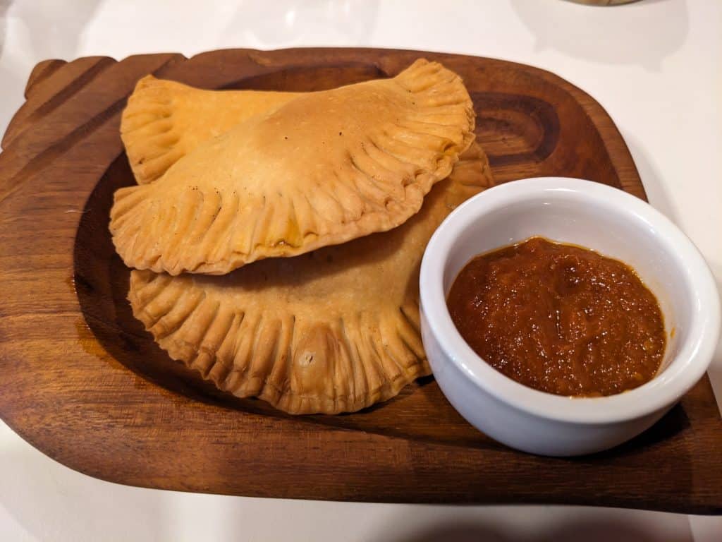 A close-up photo of two fried, flakey appetizers served on a wooden plate with a small white cup of tomato dipping sauce on the side.