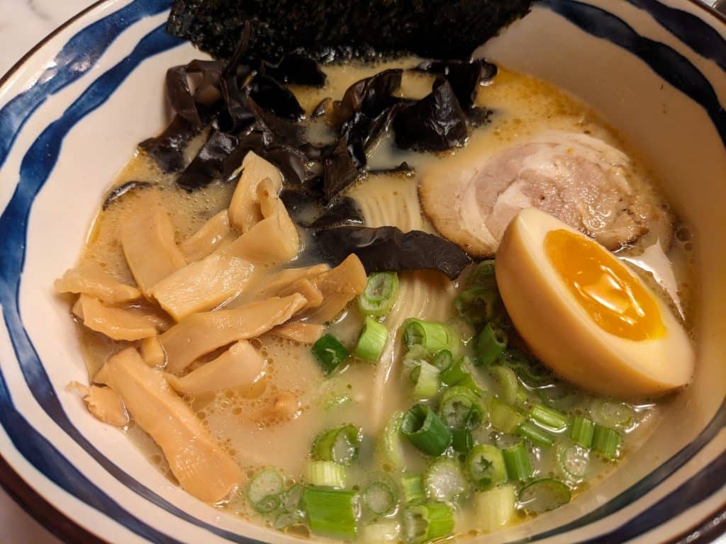 A close-up photo of a ceramic bowl of hot ramen noodles and broth. The bowl also includes half an egg, pork, and scallions.