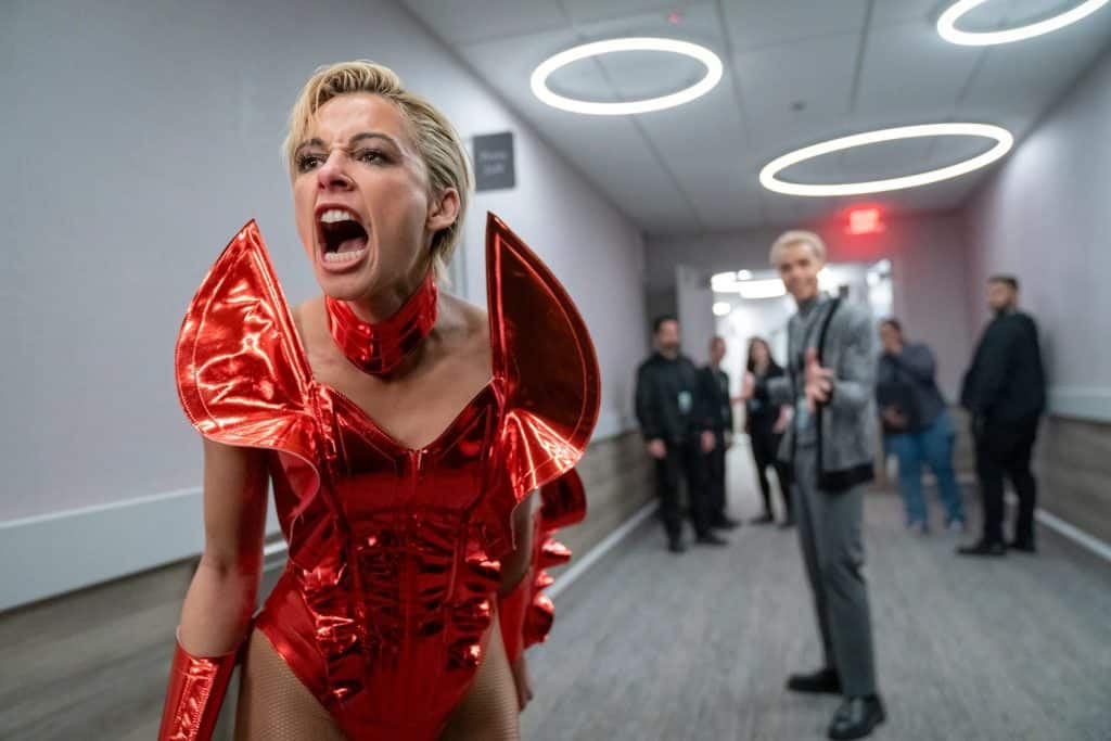 Popstar Skye Riley (Naomi Scott) shouts at someone off-screen in an elaborate red-bowlike costume in the film "Smile 2." A team peers forward towards the camera behind her, out of focus.