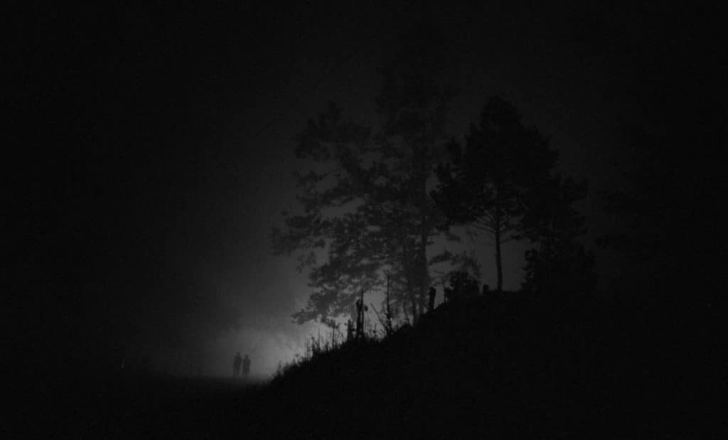 Fluorescent lights on the horizon illuminate the distant shadows of two figures during a foggy night in Southeastern Ohio.