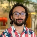 A man in his 20's with glasses, a beard, shoulder-length hair, and a red-and-white striped shirt with blue palm trees.