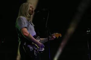 Alvvays guitarist/vocalist Molly Rankin is shwn playing a black jaguar and singing into a microphone during the band's headlining set at The Sylvee. She's wearing a light blue short-sleeved button-up and black slacks. Beams of gold light partially illuminate the backdrop behind her. She takes up the far left of the image. Apart from the lightbeams, the backdrop is black, leaving Rankin in partial shadow.