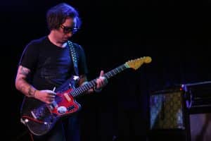 Cult Of Lip guitarist/vocalist Ronnie Lee is shown locked into concentration as he holds the whammy bar of his black Jaguar and bends a chord. He's wearing a black t-shirt and is positioned to the right of the image. A row of amps is visible in the bottom left corner. The backdrop is black.