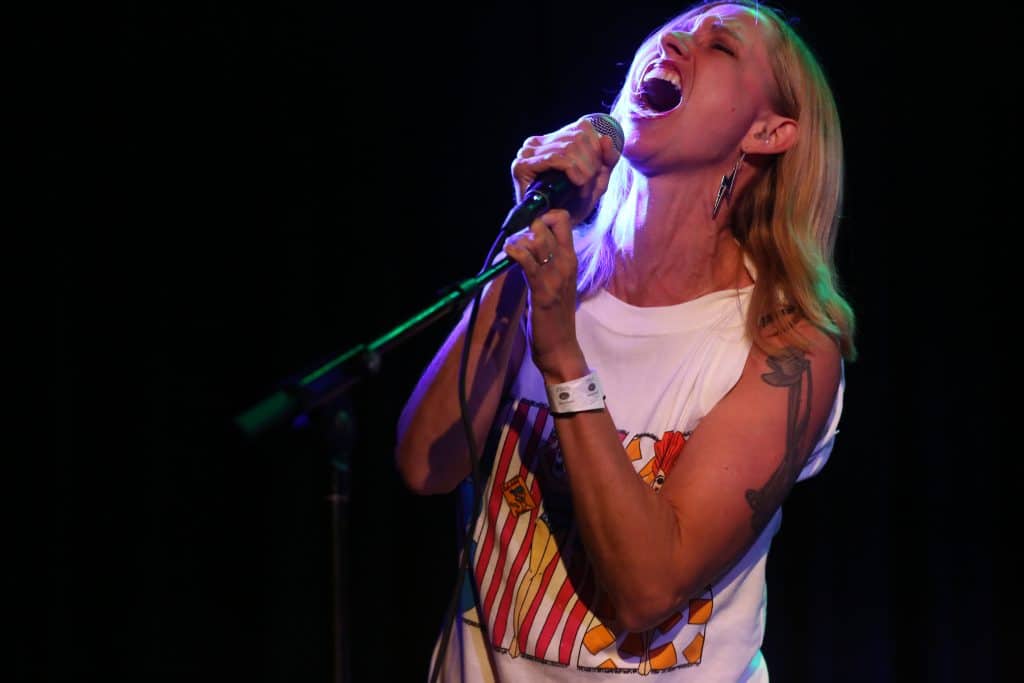 Cynthia Burnson takes a passionate guest vocal turn during Christy Costello's set at the High Noon Saloon. Burnson wears a white tank-top and appears to the right of the image, tilting her head up with her eyes firmly closed as she belts outs, with one hand wrapped around the microphone and the other on the mic stand arm. A stage light coming in from the right sharply illuminates the side of her face.