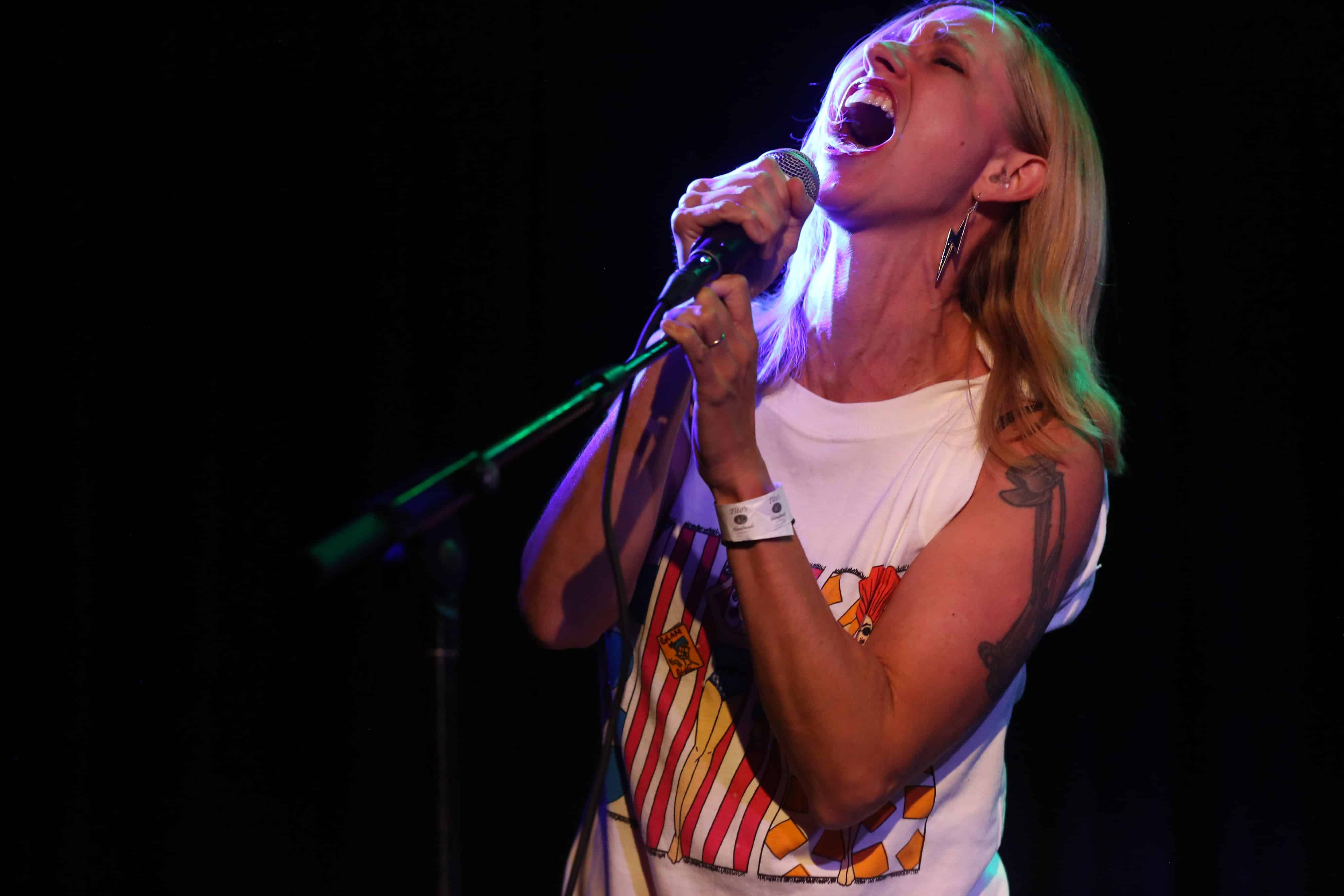 Cynthia Burnson takes a passionate guest vocal turn during Christy Costello's set at the High Noon Saloon. Burnson wears a white tank-top and appears to the right of the image, tilting her head up with her eyes firmly closed as she belts outs, with one hand wrapped around the microphone and the other on the mic stand arm. A stage light coming in from the right sharply illuminates the side of her face.