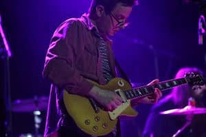 Heavy Looks' Dirk Gunderson performs during the band's opening set for The New Pornographers at The Majestic. He's wearing a horizontally striped shirt that appears green, white, and yellow, and a plum button-up. He looks down in concentration at a goldtop Epiphone Les Paul. The band's drummer is partially visible and in soft focus in the lower right corner of the image. The stage lights cast a cool blue.