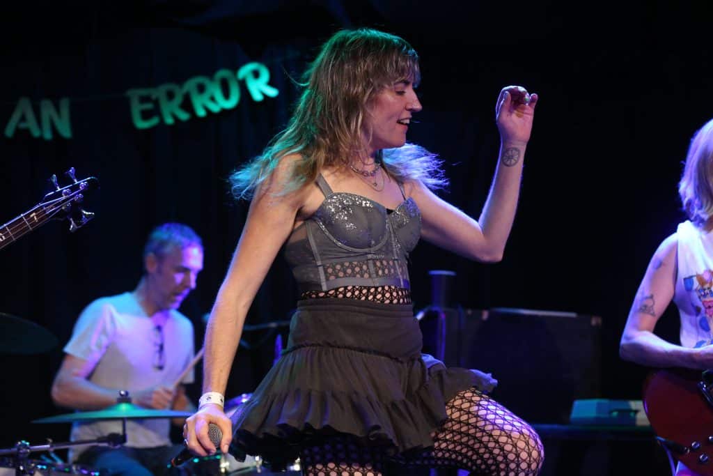 Hottt Probs perform at High Noon Saloon. Their vocalist is shown mid-strut in a black fishnet bodysuit that's worn under a black miniskirt and a gray bra with ornate designs. She takes up the middle of the image. The band's drummer is shown in soft focus in the lower left. Part of a banner cuts into the backdrop, reading "AN ERROR" behind the band.