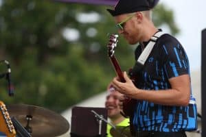 Lovely Socialite's guitarist is shown wearing cyclicst attire and neon green sunglasses while playing guitar at La Fete De Marquette. He takes up the far right of the image. The band's drummer is partially obscured in soft focus towards the bottom of the frame, just behind the guitarist, smiling. Trees are visible behind them.