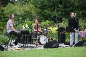 Mr. Chair play at Olbrich Botanical Gardens. Plenty of greenery is visible, as are the band's three members, who are, from left to right: seated at an electric keyboard, a drumkit, and standing in front of an upright bass. The garden that surrounds them is in bloom.