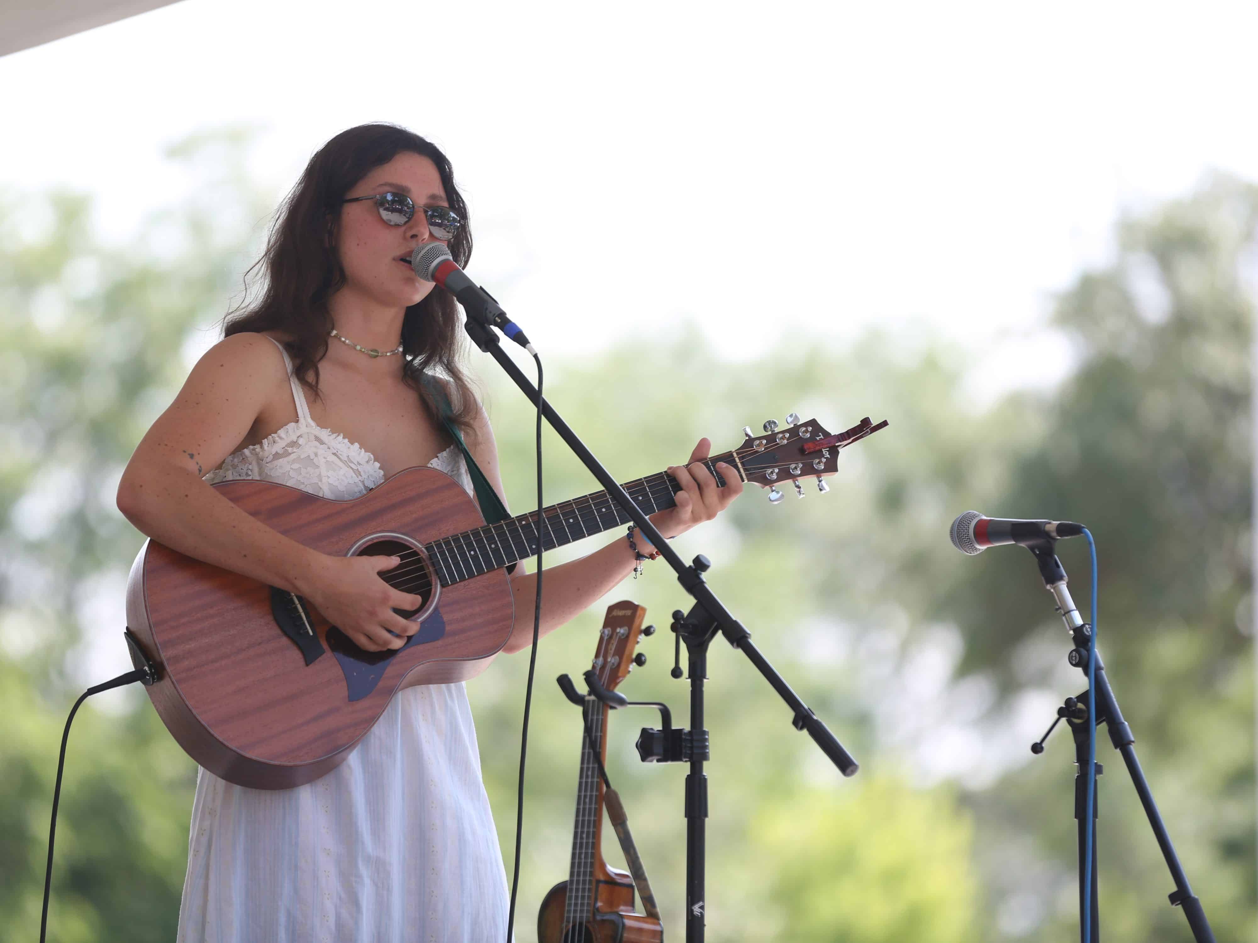 The Spine Stealers' Emma O'Shea plays a Taylor acoustic guitar and sings into a microphone during Atwood Fest in 2024. A dark red capo is clipped onto the headstock of the guitar. Oshea's wearing round sunglasses that reflect the audience in front of her, a silver choker necklace, a lace white top, and a flowing white skirt. She's positioned to the left third of the image. A pale white sky and green trees are visible behind her in the background. Directly to her side, cutting into the frame from below, is a ukulele that is mounted on a holder attached to the mic stand. An unmanned microphone is visible in the bottom right corner.