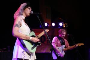 Proud Parents' C Nelson-Lifson plays telecaster and speaks into a microphone at the band's farewell show at High Noon Saloon. Nelson-Lifson is in bridal attire and playing a seagreen telecaster in the left of the image. In the bottom right, Tyler Fassnacht is visible in soft focus, holding a C chord on his guitar.