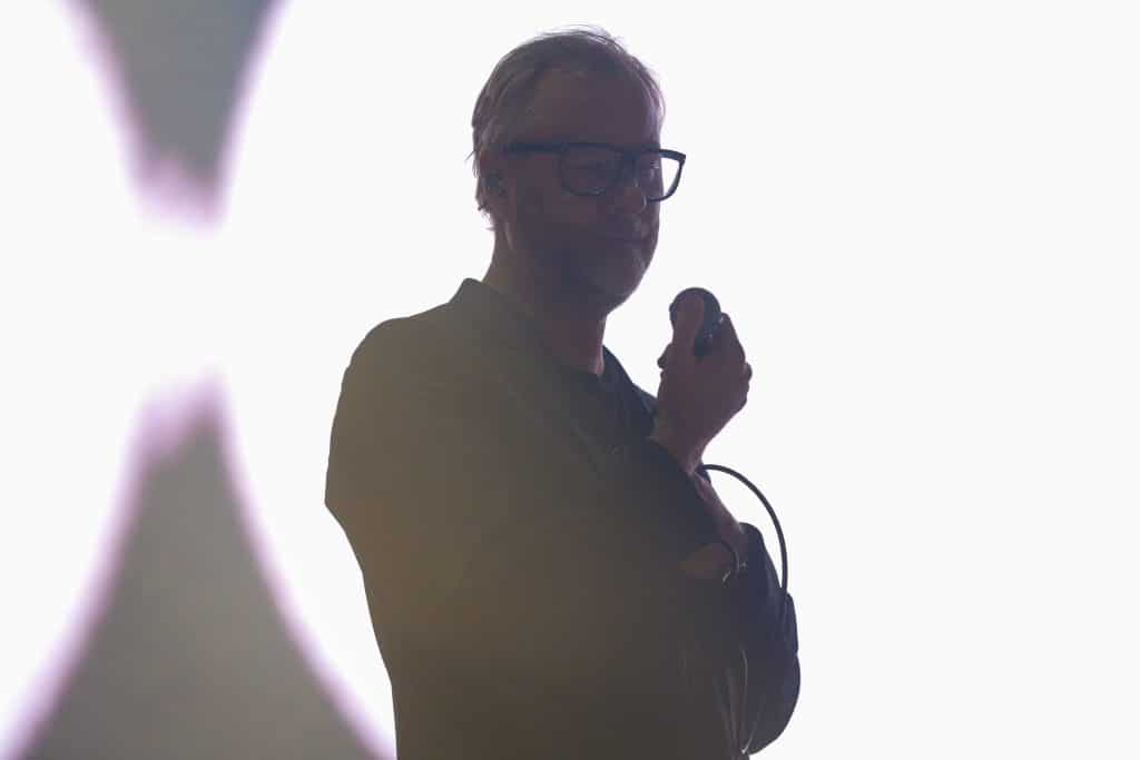 The National vocalist Matt Berninger pensively holds a microphone during the band's headlining set at Breese Stevens Field. He stands in front a blindingly white screen that washes out a good bit of color. He appears mostly in darkened silhouette, though his glasses and dark blazer can be made out.