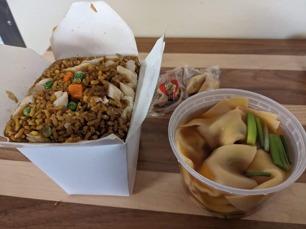 A close-up photo of two takeout containers sitting on a wooden cutting board. The white box container on the left is filled with fried rice, and the plastic cup on the right contains a dark broth with several large dumplings.