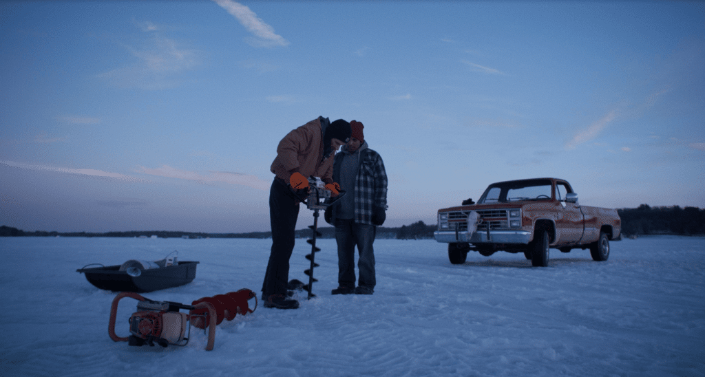 A film still captures two men standing on a snowy, frozen lake near dusk. They are bundled in heavy clothing. The older man closest to the foreground drills into the ice with an auger while the younger man watches. A red pick-up truck is parked a short distance behind them to the right.