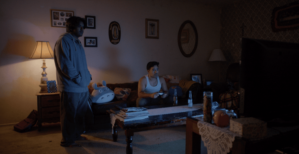 In a darkened living room lit only by a single lamp and the soft glow of the television in the right foreground, a young man stands near a couch at the left and stares at the television. The older man is seated on the couch with a game controller in hand also looking at the television.