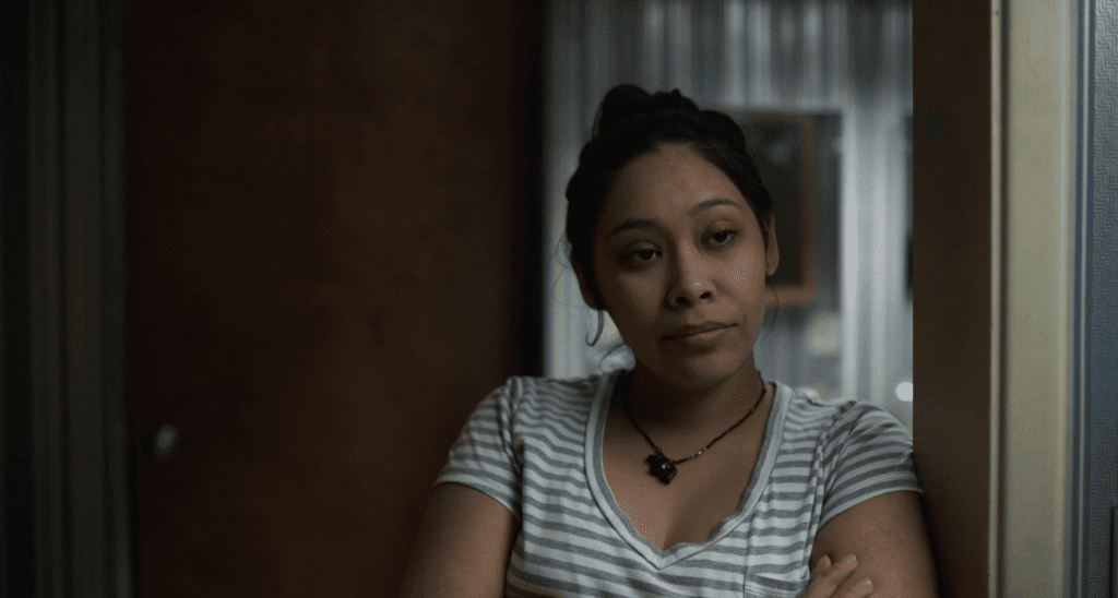 At a medium close-up, a young Hispanic woman stares forward off-screen with a slightly irked expression. Her arms appear to be folded. She wears a horizontally striped tee shirt and necklace.