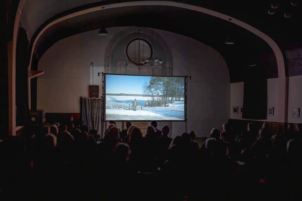 A photo at a long shot in a large, darkened room captures a sizable audience seated and staring forward at an illuminated projection screen. The screen displays a snowy still from the film "February."