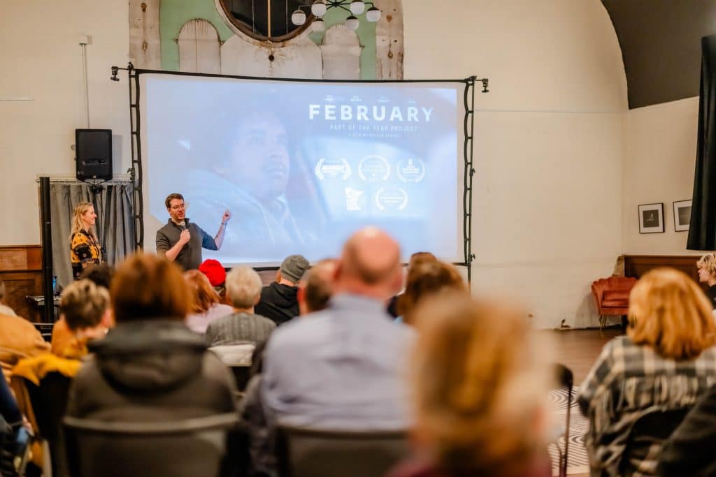 In a large, brightly lit room, several people sit and stare forward at a man holding a microphone and talking in front of a projection screen.