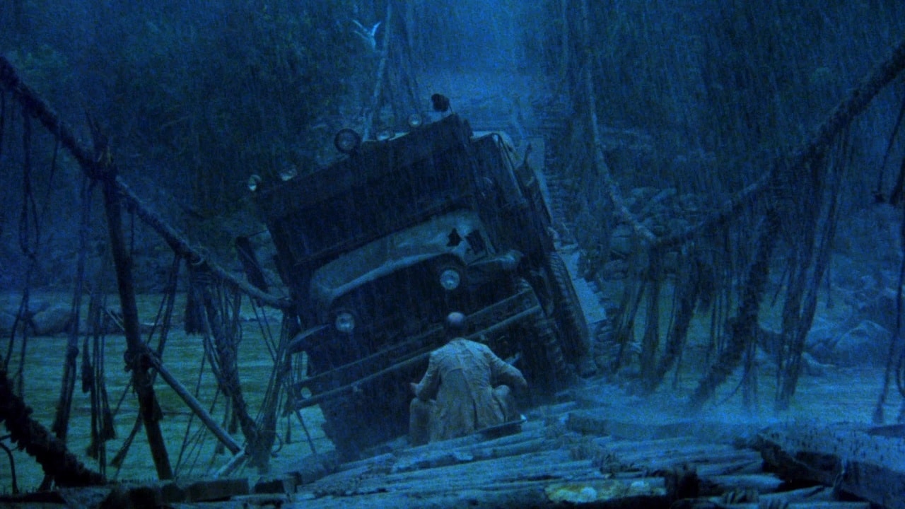 A blue-tinted image of an unsteady bridge with a tanker truck in the middle, tilting to one side as if nearly collapsing the ropes and planks due to its weight. A man squats awkwardly in front of the truck with his back to the camera.