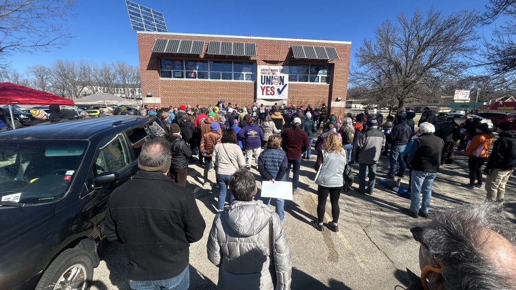 A photo shows a crowd of people at a labor rally, seen from the back/