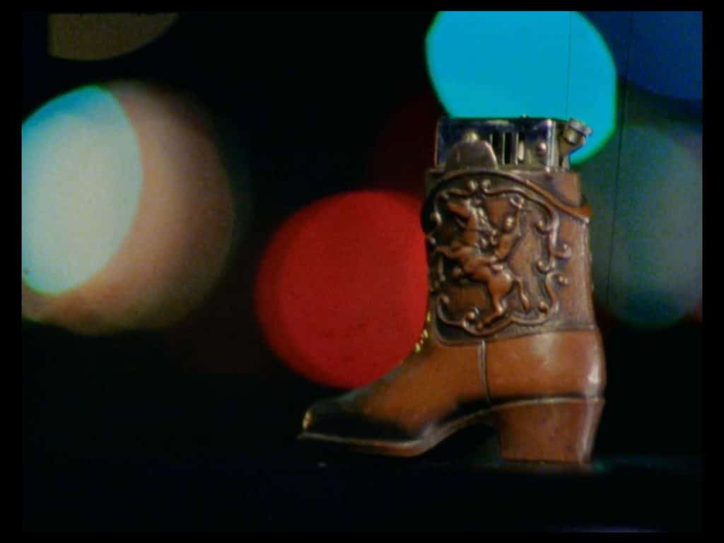 A still from the film Milk Punch shows a close-up of a novelty lighter shaped like a cowboy boot. In the background, the night sky and blurred traffic lights are visible.