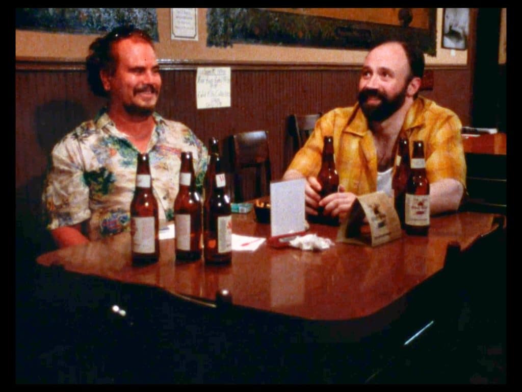 A still from the film Milk Punch shows two men seated at a table strewn with beer bottles. They are smiling. One is wearing a loud Hawaiian shit, the other a more modest yellow checked shirt.