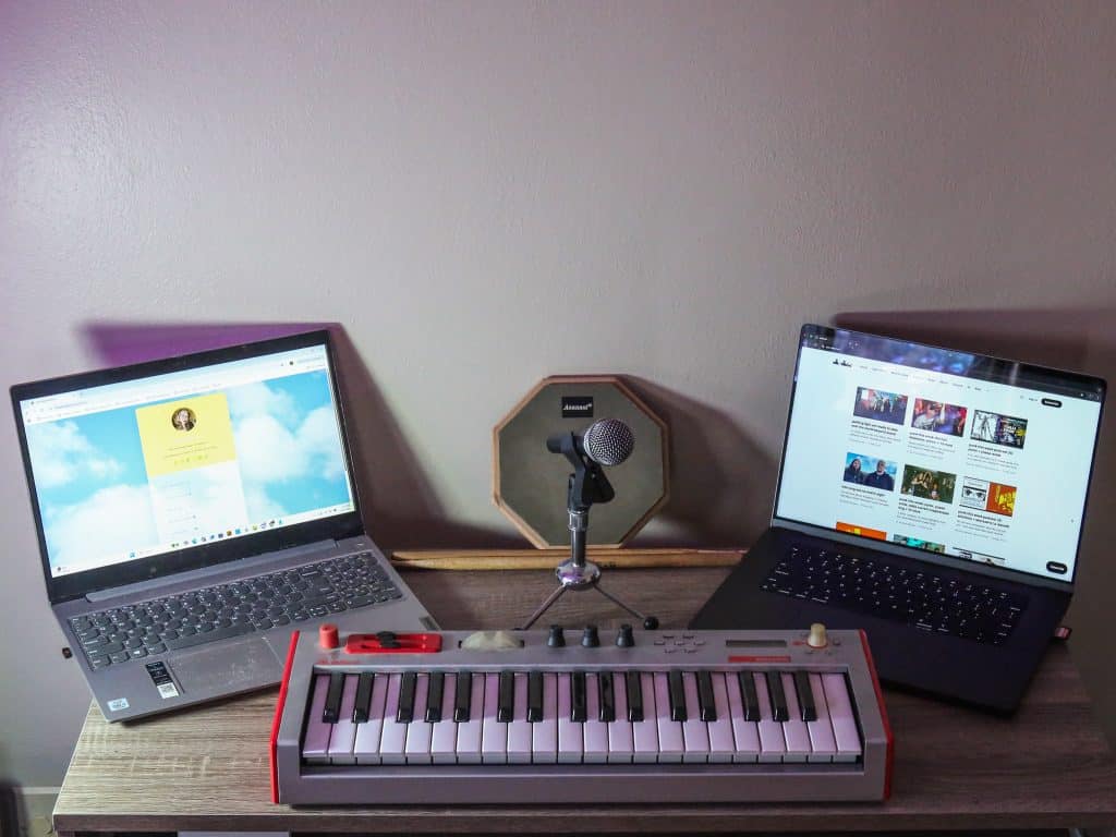Two laptops are open on either side of a desk that's pushed against a white wall. The laptop on the left is open to the home page of The Yellow Button, the laptop on the right is open to the home page of see/saw. Between the laptops, from closest to the camera to furthest away, are a synth, a microphone clamped into a small stand, and two drumsticks in front of a practice drum pad.