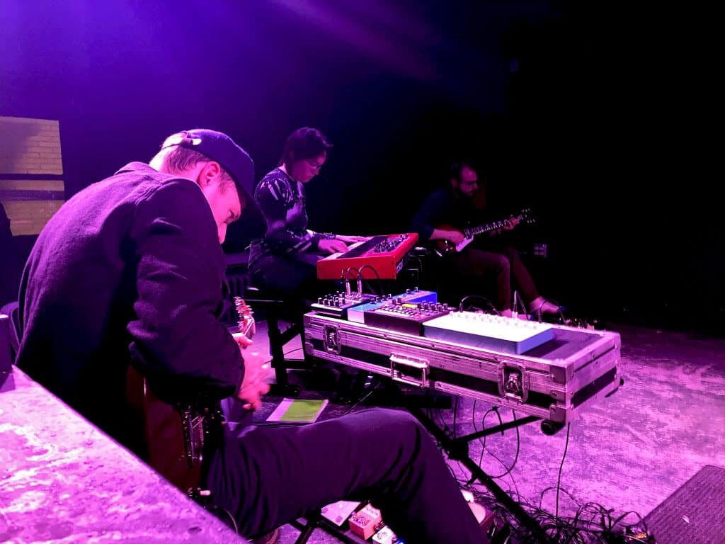 From left to right: Andrew Fitzpatrick, Emili Earhart, and Julian Lynch. Fitzpatrick performs with Earhart and Lynch during his Observatory series at Gamma Ray Bar. Fitzpatrick is wearing dark clothing anda dark baseball cap, hunched over on a stool playing guitar with a row of synthesizers directly in front of him. Earhart's also seated, playing piano. Lynch is also sitting, playing guitar, and maneuvering his feet over some effects pedals in front of him.