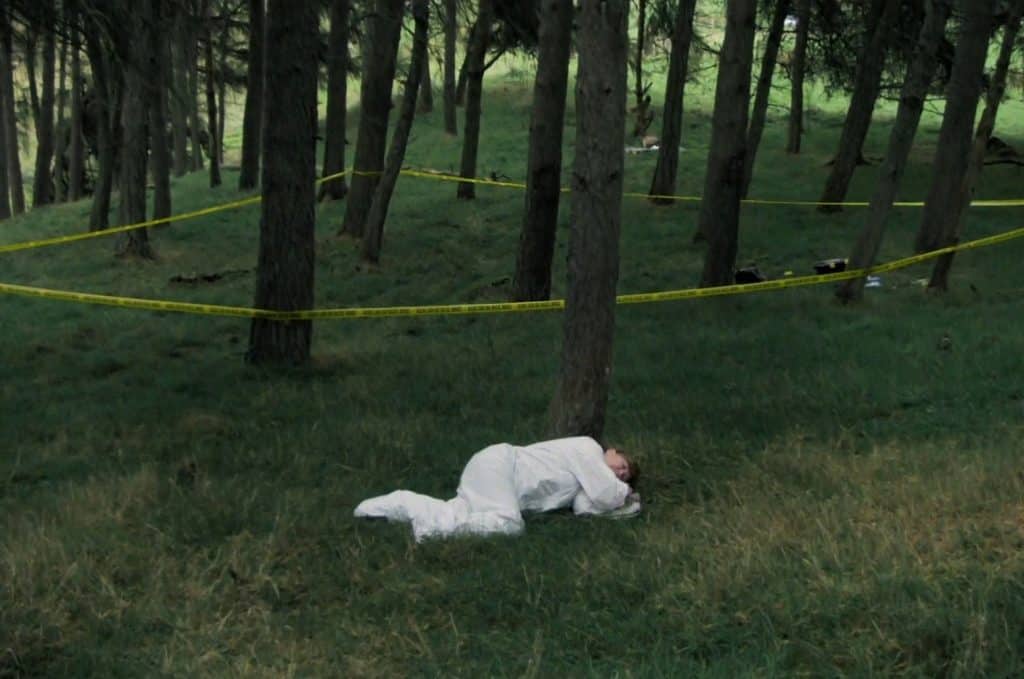 In the foreground of an image in a forested area, a woman in a white lab suit lays on the grass in the fetal position. Behind her, yellow tape that faintly reads "crime scene do not enter" is wrapped in a semi-triangular shape around a cluster of trees.