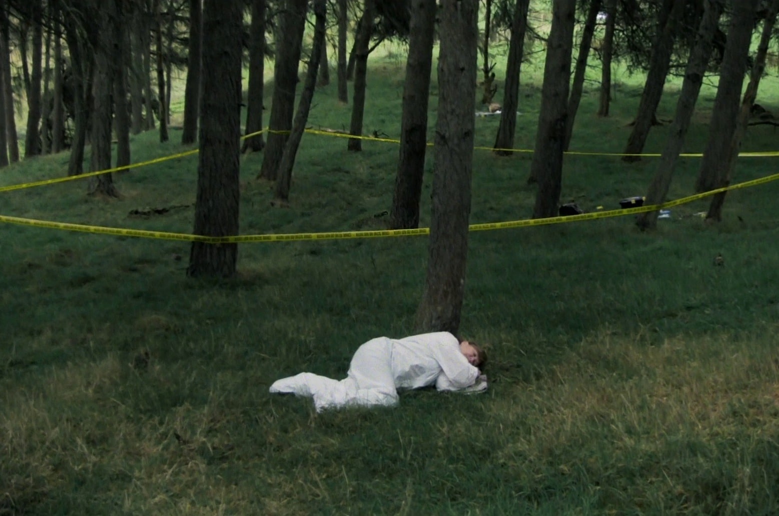 In the foreground of an image in a forested area, a woman in a white lab suit lays on the grass in the fetal position. Behind her, yellow tape that faintly reads "crime scene do not enter" is wrapped in a semi-triangular shape around a cluster of trees.