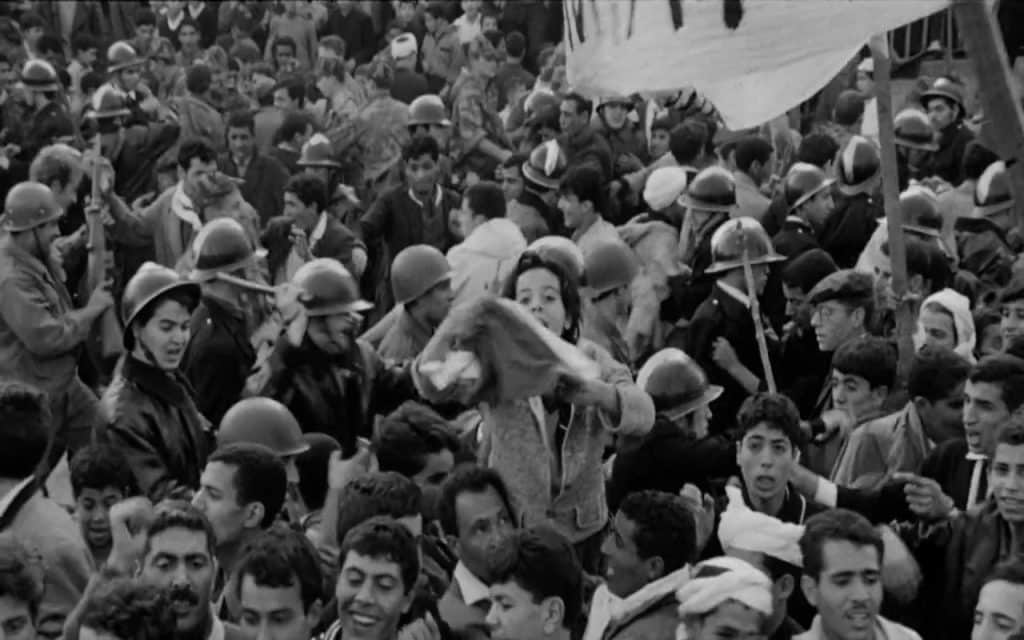 A mid-motion still frame shows dozens of soldiers and civilians who've gathered outside in a demonstration. Centered in the frame is a young person reaching towards the camera and staring directly into the lens; they're waving what looks to be a cloth flag.
