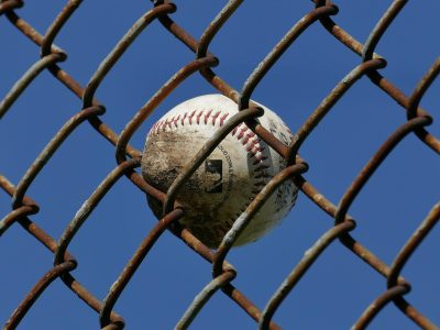 A close-up of a dirtied, official Major League Baseball baseball stuck in a rusty chain-link fence against a cloudless blue sky.