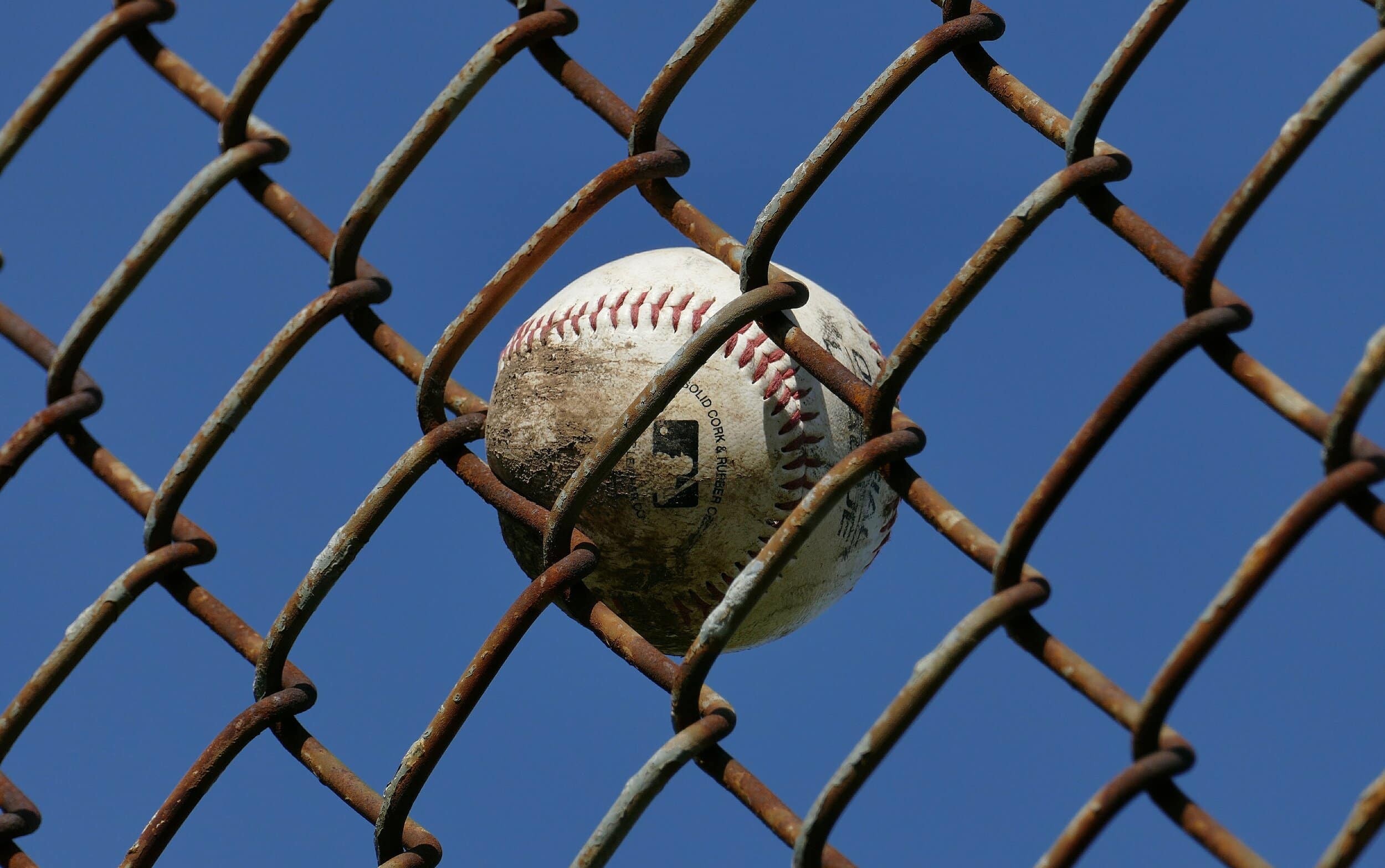 A close-up of a dirtied, official Major League Baseball baseball stuck in a rusty chain-link fence against a cloudless blue sky.