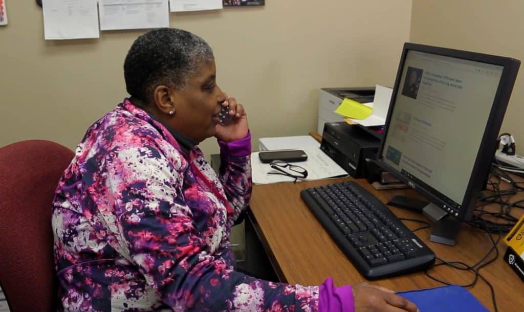 A frame from the short film "Choose The Pond" shows a middle-aged woman wearing a long-sleeve purple and red tie-dye shirt. She sits in profile at a desk in a corner, and looks ahead at a computer monitor, while speaking on the phone.