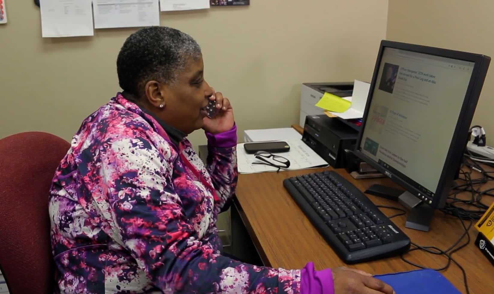 A frame from the short film "Choose The Pond" shows a middle-aged woman wearing a long-sleeve purple and red tie-dye shirt. She sits in profile at a desk in a corner, and looks ahead at a computer monitor, while speaking on the phone.