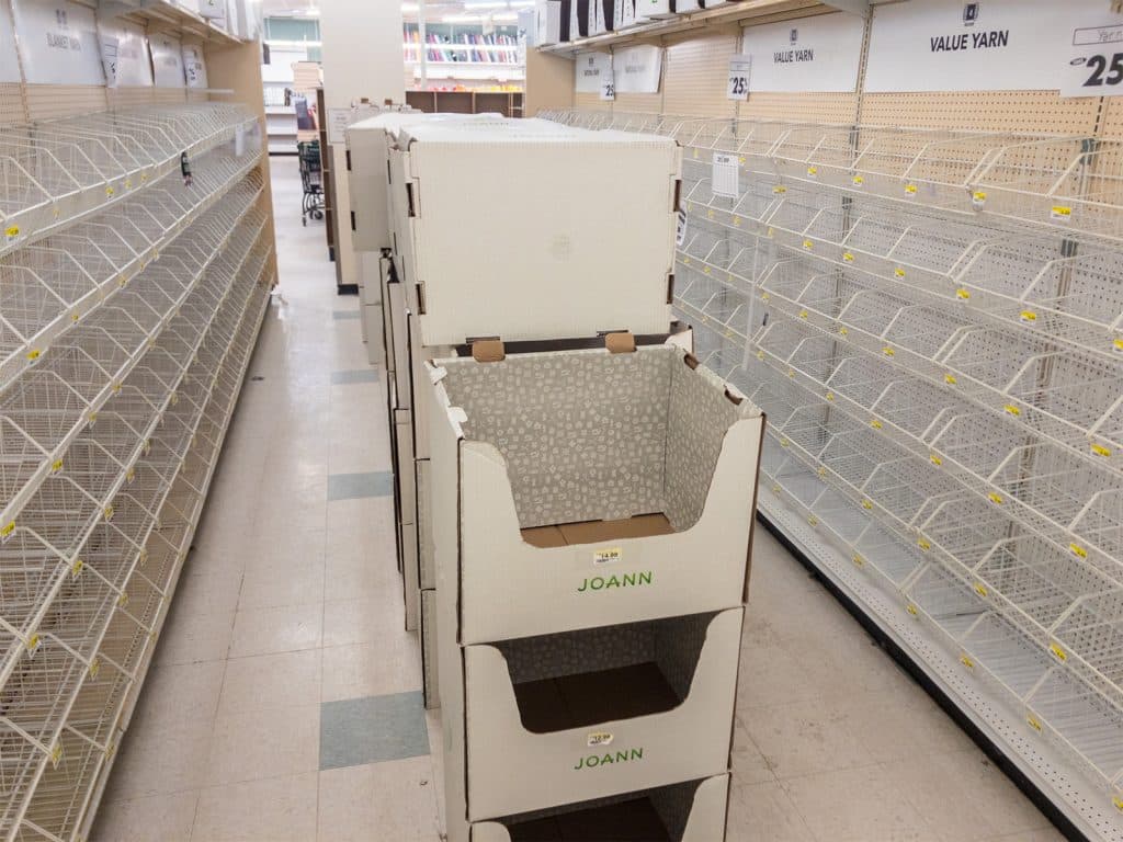 A photo shows an aisle at a closing Joann Fabrics store with an empty expanse of product racks. Several white rectangular signs at the top of the shelves on the right-hand side read "VALUE YARN" in black lettering.