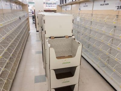 A photo shows an aisle at a closing Joann Fabrics store with an empty expanse of product racks. Several white rectangular signs at the top of the shelves on the right-hand side read "VALUE YARN" in black lettering.