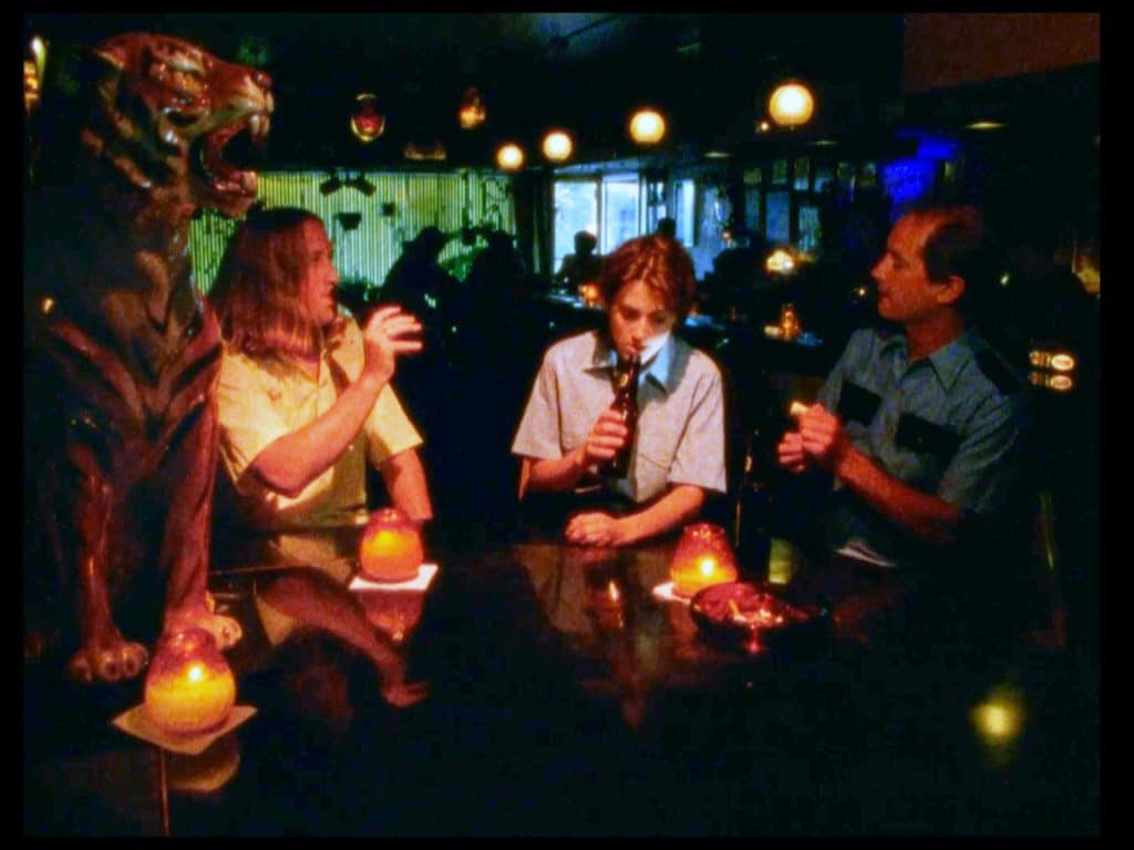 A still from the film Milk Punch shows three characters seated at a table in the back of a dark bar, Le Tigre Lounge. In the left foreground is one of the ceramic tiger statues decorating the bar.