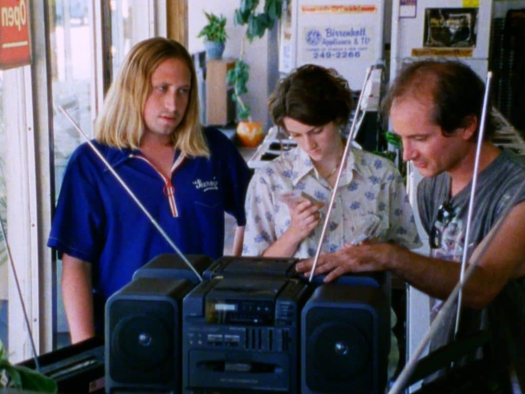 A still frame from the film "Milk Punch" shows three characters in their 20s standing near the windows of an appliance store in daylight. They hover around a stereo boombox in the foreground. The balding man on the right puts his left hand on top of it, inspecting it, while the woman in the center inspects a cassette tape case. To the left, the other long-haired man stares with an derisive expression.
