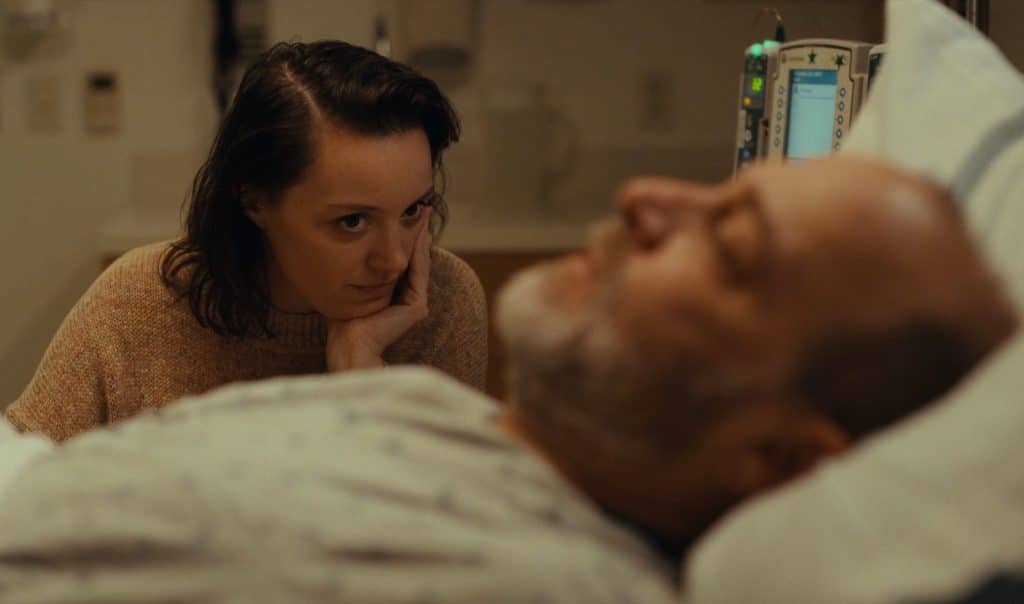 A color-muted image shows a middle-aged male hospital patient laying on a bed out of focus. Behind him is a younger woman sitting in the middleground of the frame. She is in focus, and stares affectionately at him with her left hand cradling her chin.