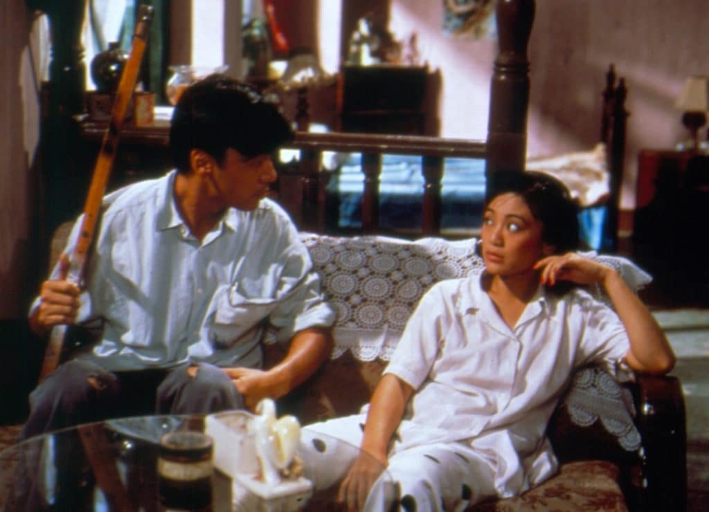 A still image from the film "Shanghai Blues" shows a young couple sitting somewhat anxiously on a couch that's in front of a bedroom area behind them. They stare at each other intently with straight faces. The woman on the right's left arm is resting on the arm of the couch, while the man on the left is holding a thin, elongated wooden prop or piece of a wooden frame that's resting on the floor.