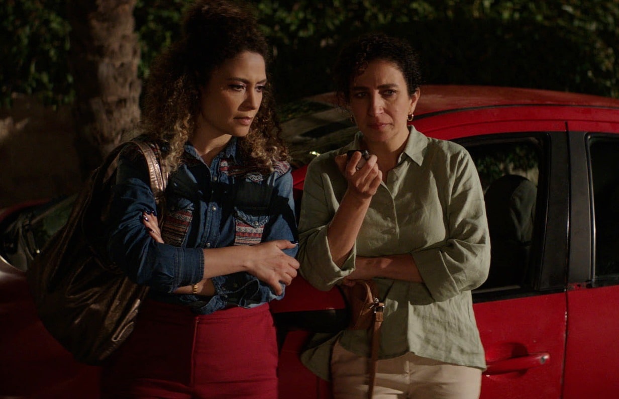 Two woman stand in front of a red parked car at night outside. The woman on the right holds her phone up with her right hand, closer to her face, while the other woman leans in slightly with her arms folded. Both women stare ahead with calm, neutral expressions that suggest they are listening attentively.
