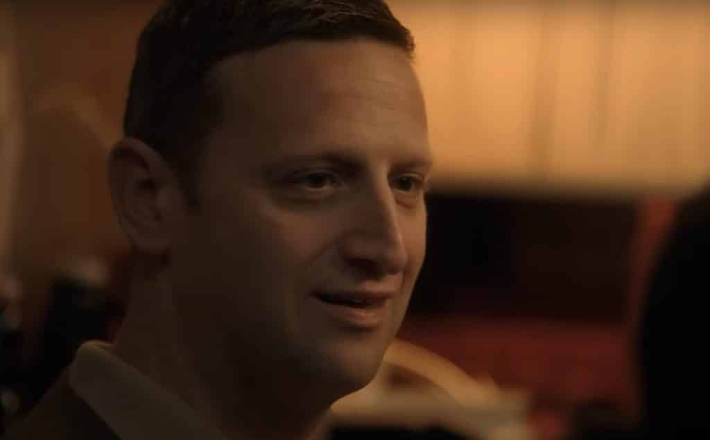 A close-up and rather dimly lit shot of a man's face turning to the right to gaze off screen. His expression is awkwardly deadpan and mannequin-like, cracking a slight smile.