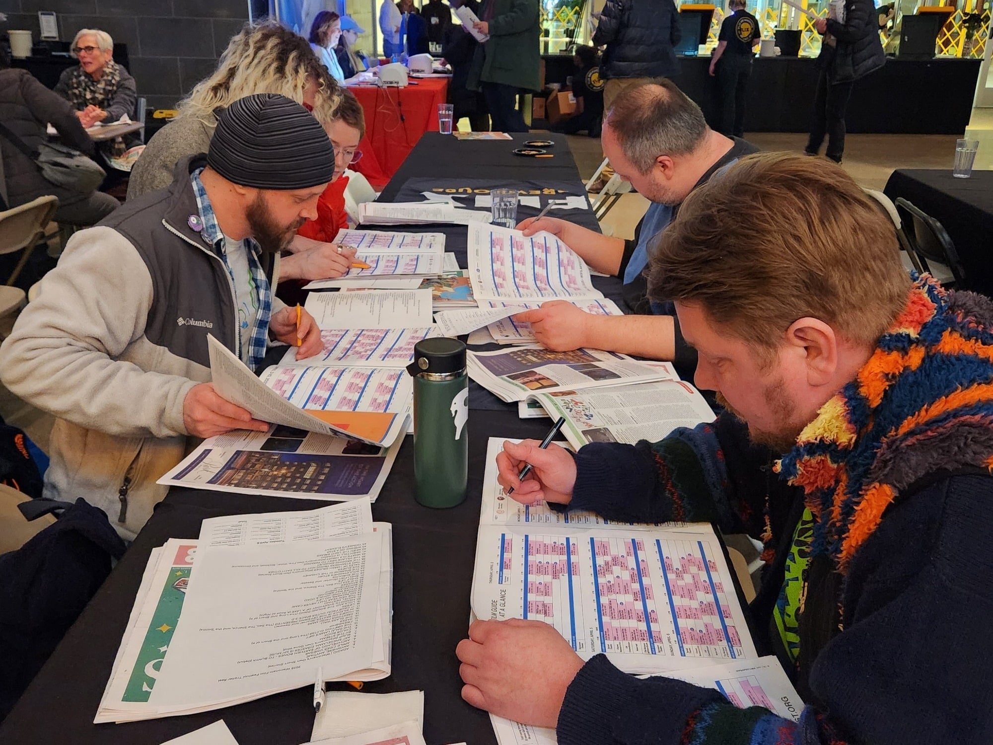 Five people sit at an elongated table in a moviehouse lobby. Two are seated on the right side, closer to or in the foreground, while the three on the left sit further away in the middle ground. All of them carefully pore over printed materials on the table. Other attendees mill about in the background.