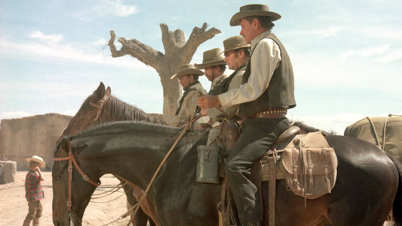 In a dusty, desert landscape, four cowboys sit on horses in an askew vertical row extending from the foreground into the middleground of the frame so that each of their bodies is visible. Each man stares down at something in their hands.
